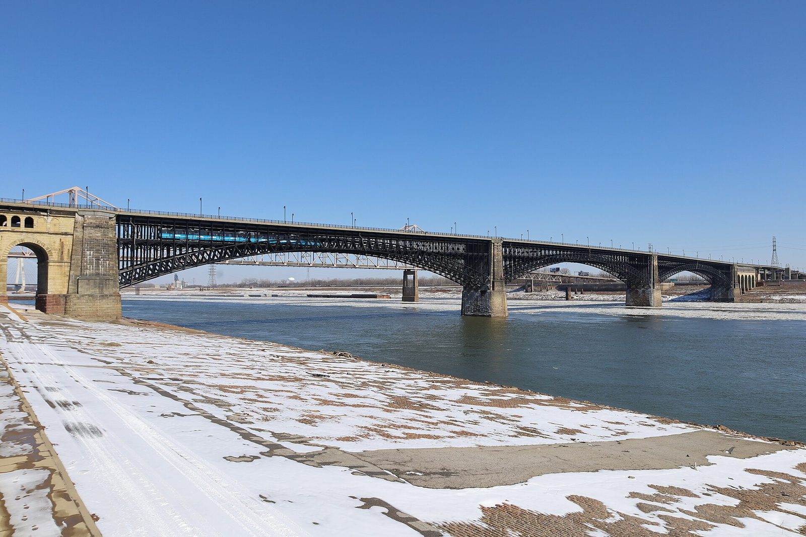 The Eads Bridge spanning the Mississippi River at St. Louis under a clear winter sky, with a MetroLink train crossing and snow on the levee