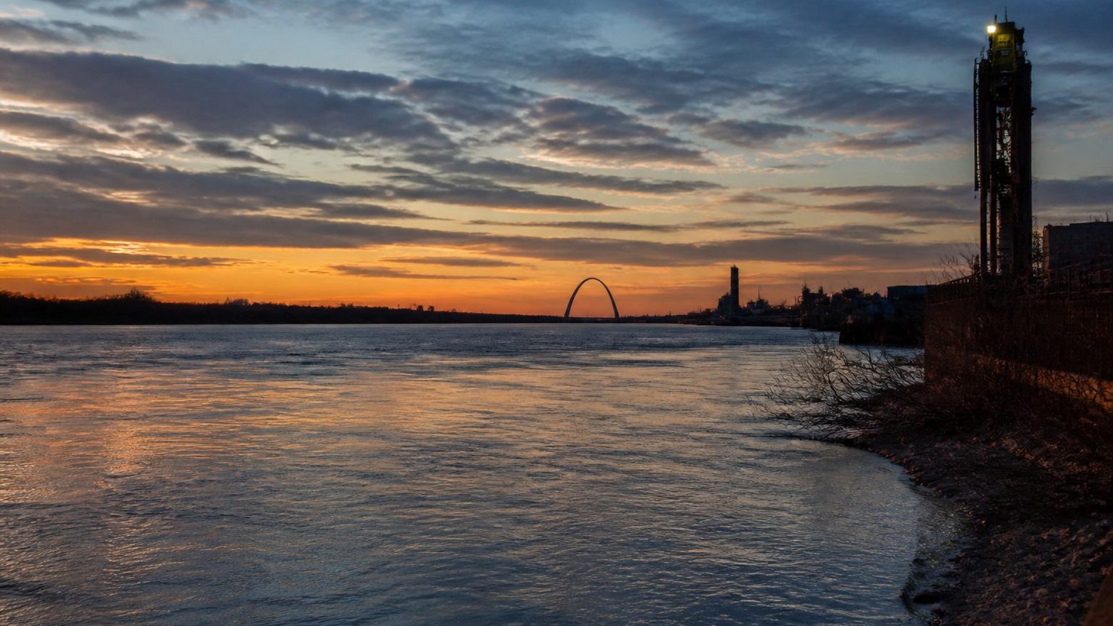 The Gateway Arch on the horizon at sunset, seen across the wide Mississippi River from the riverbank